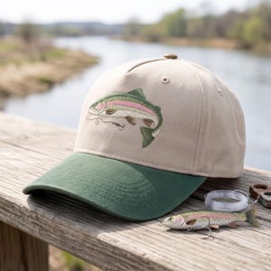 Op de afbeelding: Een beige honkbalpet met een donkergroene klep en een geborduurde afbeelding van een regenboogforel op het voorpaneel. De forel is afgebeeld in tinten groen, roze en wit, met een vishaak en vislijn zichtbaar onder de vis.