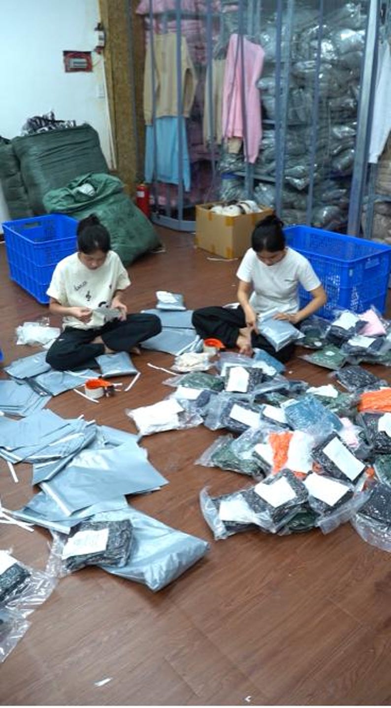 May include: Two individuals in white t-shirts and dark pants sit on a wooden floor, surrounded by numerous plastic-wrapped clothing items and shipping mailers. Blue plastic crates and racks filled with folded garments are visible in the background.