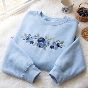 Op de afbeelding: Een lichtblauwe sweater met ronde hals en een geborduurd ontwerp van donkerblauwe bosbessen en lichtgroene bladeren op de borst. Het borduurwerk toont trossen ronde bessen met gedetailleerde schaduwen en bladeren in een horizontale opstelling.