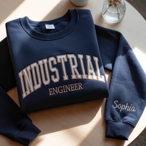 May include: A navy blue sweatshirt featuring the embroidered text "INDUSTRIAL ENGINEER" in a cream-coloured, collegiate font. The name "Sophia" is embroidered on the sleeve. The garment is displayed on a wooden surface.