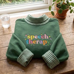 May include: A forest green sweatshirt featuring a mock neck collar and sleeve cuffs with a green and white gingham pattern. The chest displays the embroidered text "speech therapy" in multicoloured letters surrounded by small embroidered star accents.