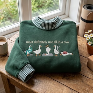 Könnte beinhalten: Ein dunkelgrünes Sweatshirt mit Stehkragen und grün-weißem Vichy-Muster. Auf der Vorderseite befindet sich eine Stickerei von fünf Gänsen in verschiedenen Posen mit dem Text, most definitely not all in a row.