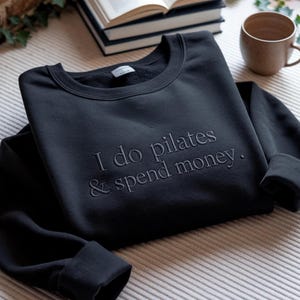May include: Black crewneck sweatshirt with the embroidered text "I do pilates & spend money." The sweatshirt is folded on a patterned surface, with a book and a mug in the background.