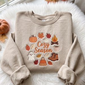Op de afbeelding: Een beige sweater met ronde hals voorzien van geborduurde herfstthema-iconen, waaronder een pompoen, kampvuur, spookje, mok, laarzen, muts en diverse bladeren rondom de oranje tekst die Cozy Season spelt in een cursief lettertype.