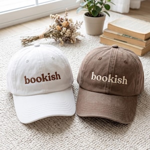May include: Two baseball caps, one white with brown embroidered text reading bookish and one brown with white embroidered text reading bookish. Each cap features a curved brim, a button on the top, and stitched eyelets for ventilation.