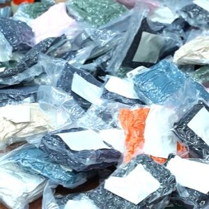 May include: A large collection of assorted textile items individually packaged in clear plastic bags. Each bag features a white rectangular label on the front. The items vary in color, including shades of black, blue, green, orange, pink, and beige.