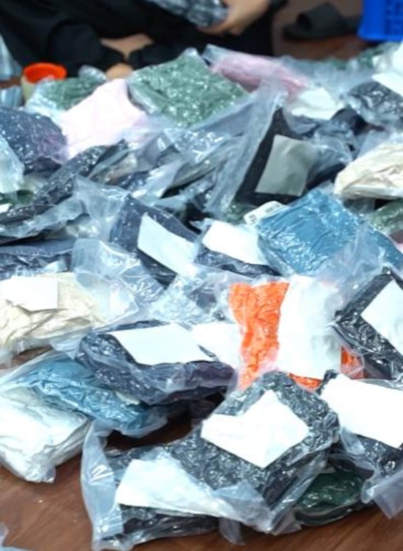 May include: A large collection of assorted textile items individually packaged in clear plastic bags. Each bag features a white rectangular label on the front. The items vary in color, including shades of black, blue, green, orange, pink, and beige.