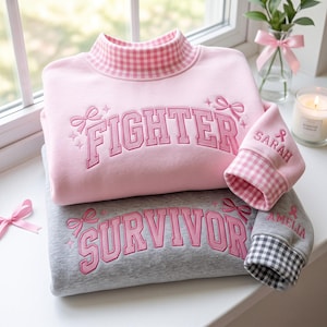 Op de afbeelding: Een lichtroze sweatshirt met een opstaande kraag en vichy-patroon, voorzien van de geborduurde tekst FIGHTER en een strik. Een grijs sweatshirt met de tekst SURVIVOR en een strik. Beide hebben vichy-boorden en namen.