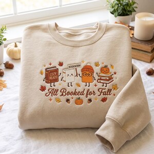 Puede incluir: Una sudadera de cuello redondo en color beige con un diseño bordado de cuatro libros antropomórficos con caras, incluyendo uno con gorro y gafas, rodeados de hojas de otoño, bellotas y una calabaza, con el texto All Booked for Fall.