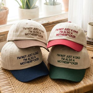 May include: Four baseball caps with cream-colored crowns and contrasting brims in brown, red, navy blue, and dark green. Each cap features embroidered text that reads: I'M NOT JUST A DAD I'M ALSO A MOTHERF*CKER in matching colors to the brims.