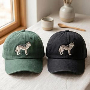 May include: Two baseball caps, one in olive green and one in black, each featuring an embroidered graphic of a standing gray wolf on the front panel. Both caps have a structured crown, a curved brim, and a matching button on the top center.