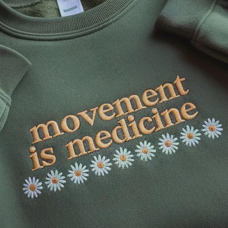 Pu&ograve; includere: Una felpa verde oliva con la scritta ricamata "movement is medicine" in oro. Sotto il testo, una fila di margherite ricamate con petali bianchi e centri gialli.