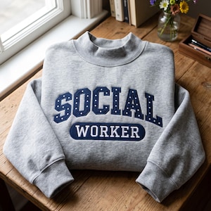 May include: A heather grey mock neck sweatshirt featuring the text SOCIAL WORKER in dark blue embroidered lettering. The word SOCIAL is arched in a collegiate font with small white dots, while WORKER is in a solid dark blue oval patch.