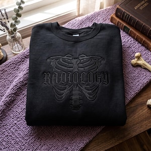 May include: A black crewneck sweatshirt featuring a tonal black embroidered design of a human rib cage and spine. The word RADIOLOGY is embroidered in a gothic-style font across the center of the rib cage graphic on the front of the garment.