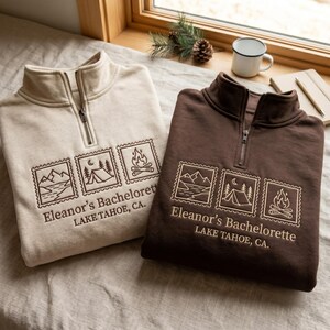 May include: A cream and a dark brown quarter-zip sweatshirt, each featuring embroidered stamp-style graphics of mountains, a tent, and a campfire. Below the graphics, the text reads: Eleanor's Bachelorette LAKE TAHOE, CA. Both garments have a high collar.