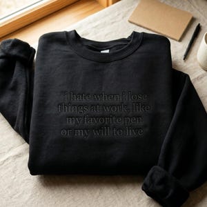 Könnte beinhalten: Ein schwarzes Sweatshirt mit Rundhalsausschnitt und tonaler schwarzer Stickerei auf der Brust. Der Text lautet: i hate when i lose things at work, like my favorite pen or my will to live. Das Kleidungsstück hat lange Ärmel mit Rippbündchen.