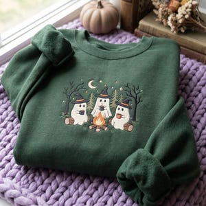 May include: A dark forest green crewneck sweatshirt featuring an embroidered design of three white ghosts wearing black witch hats. The ghosts sit on logs around a small campfire, holding mugs, surrounded by trees, a crescent moon, and stars.