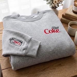 以下が含まれることがあります： 胸元に赤と黒のDiet Cokeロゴが刺繍された、ヘザーグレーのクルーネックトレーナー。袖口には、赤い文字でブランド名が記されたDiet Cokeの缶の小さな刺繍グラフィックが施されています。