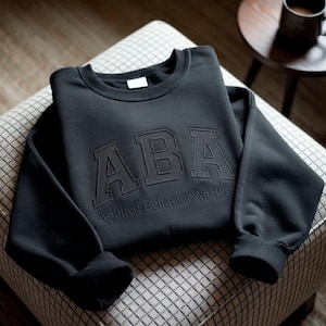 May include: A black crewneck sweatshirt featuring the letters ABA in a large, textured, collegiate-style font across the chest. Below the letters, the text Applied Behavior Analysis is embroidered in a smaller, matching black font.