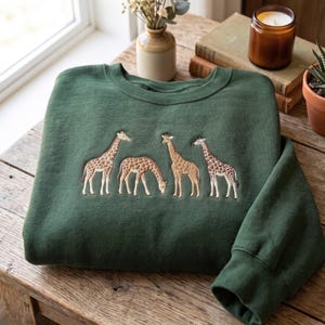 Puede incluir: Una sudadera de cuello redondo en color verde bosque con un diseño bordado de cuatro jirafas en varias posturas. Las jirafas están representadas en tonos marrones y bronceados, mostrando sus patrones de manchas distintivos en el centro.