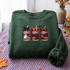 May include: A dark green crewneck sweatshirt featuring an embroidered design of three soda cans labeled Dr Pepper. Each can is decorated with a bow and Halloween motifs including a ghost, a pumpkin, a haunted house, a crescent moon, and bats.