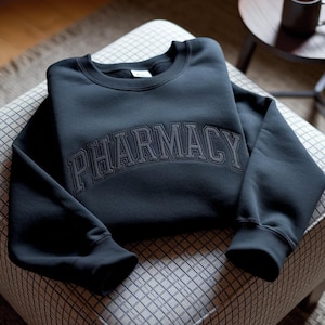 May include: A dark grey crew neck sweatshirt with the word "PHARMACY" embroidered across the chest in a darker grey, arched font. The sweatshirt is folded on a patterned ottoman. The sleeves are cuffed at the wrists.