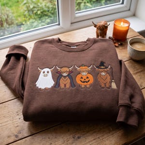 Könnte beinhalten: Ein braunes Sweatshirt mit Rundhalsausschnitt und einer Stickerei von vier Highland-Rindern in Halloween-Kostümen: eines als Geist, eines als Vampir, eines in einem Kürbis und eines als Hexe mit einem Besen.