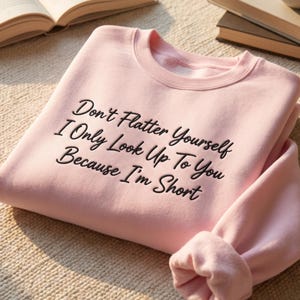 Pode incluir: Camisola rosa claro com a frase bordada "Don't Flatter Yourself I Only Look Up To You Because I'm Short". Tem gola redonda e mangas compridas. O tecido parece macio, possivelmente uma mistura de algodão.