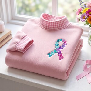 Op de afbeelding: Een lichtroze sweatshirt met een opstaande kraag en mouwboorden in een roze-wit vichy-ruitpatroon. De voorzijde van het kledingstuk toont een geborduurd bewustzijnslintje, samengesteld uit kleine, veelkleurige hartjes in tinten teal, paars en magenta.