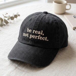 Könnte beinhalten: Eine schwarze Baseballkappe mit verwaschener Textur und dem gestickten Text 'be real, not perfect.' in hellem Beige. Die Kappe hat einen gebogenen Schirm, einen Knopf oben in der Mitte und genähte Ösen zur Belüftung an der Kappe.
