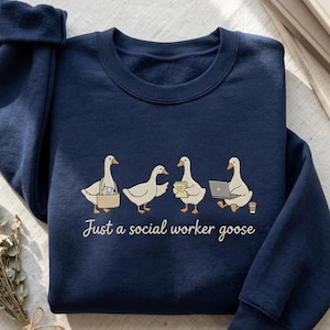 Puede incluir: Sudadera azul marino con un diseño de gansos de dibujos animados, cada uno sosteniendo objetos relacionados con el trabajo como un maletín, libros, un portátil y una taza de café. El texto "Just a social worker goose" está impreso debajo.