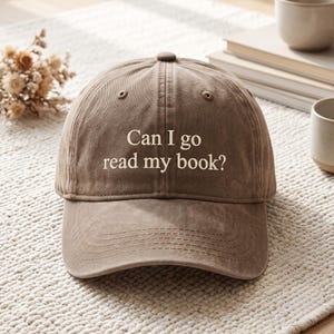 Könnte beinhalten: Eine braune Baseballkappe mit sechs Paneelen und einem gebogenen Schirm. Auf der Vorderseite der Kappe befindet sich der gestickte Text "Can I go read my book?" in einer hellen Cremefarbe. Der Stoff hat eine leicht verwaschene, matte Textur.