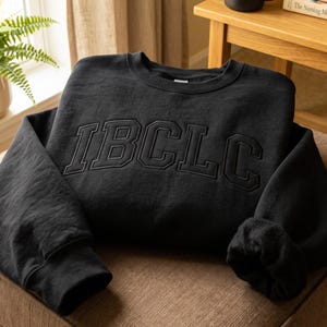 May include: A black crewneck sweatshirt featuring the letters IBCLC embroidered in a matching black thread across the chest. The garment has long sleeves with ribbed cuffs and a ribbed neckline, showcasing a clean, monochromatic design.