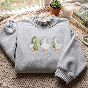 Könnte beinhalten: Ein hellgraues Sweatshirt mit Stehkragen und Bündchen in weiß-grauem Vichy-Karo-Muster. In der Mitte des Sweatshirts befindet sich eine Stickerei mit drei weißen Enten, von denen eine ein Gurkenkostüm trägt, eine eine Gurke hält und eine ein Glas hält.