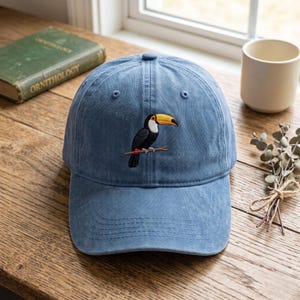 May include: A blue baseball cap featuring an embroidered toucan perched on a branch. The bird has a black body, a white chest, and a large yellow and orange beak. The cap has a structured crown, a curved brim, and a button on the top center.