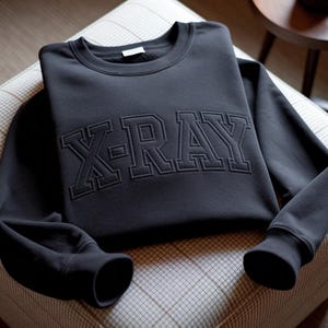 May include: A black sweatshirt featuring the word "X-RAY" embroidered in large, block letters. The garment is neatly folded, displaying the textured embroidery. The sleeves have ribbed cuffs. The sweatshirt is displayed on a patterned surface.