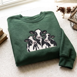 Könnte beinhalten: Ein dunkelwaldgrüner Rundhals-Pullover mit einer zentralen Stickerei von drei schwarz-weißen Kühen. Das Design zeigt die Köpfe der Kühe, die eng beieinander positioniert sind und detaillierte Gesichtszüge sowie Fellzeichnungen aufweisen.