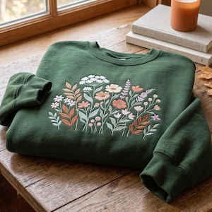 May include: A dark forest green crewneck sweatshirt featuring an embroidered design of various wildflowers and foliage in shades of white, soft pink, peach, and muted orange, centred on the front chest with long sleeves and ribbed cuffs.