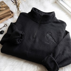 Peut inclure: Un sweat-shirt noir à fermeture éclair quart de tour avec une broderie détaillée représentant un corbeau sur la poitrine gauche. Le vêtement présente un col montant, une fermeture zippée centrale et des manches longues à poignets côtelés.