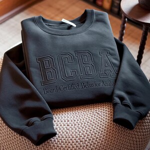 Może przedstawiać: Ciemnoszara bluza z haftowanymi literami "BCBA" z przodu oraz słowami "Board Certified Behavior Analyst". Bluza jest złożona i spoczywa na tkanej powierzchni.