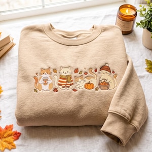 Pode incluir: Uma camisola bege de gola redonda com um design bordado de quatro gatos. Um gato segura uma chávena de café, um usa uma camisola às riscas e um cachecol, um está deitado perto de uma abóbora e um usa um gorro com um cesto de abóboras.