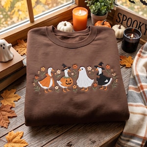 May include: A brown crewneck sweatshirt featuring an embroidered design of four white geese dressed in Halloween costumes, including a pumpkin, a ghost sheet, and witch hats, surrounded by small orange and white flowers and green foliage.