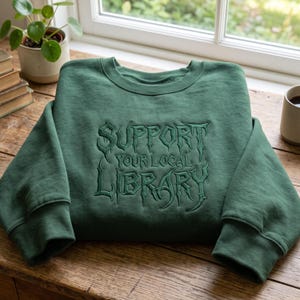 Puede incluir: Una sudadera de cuello redondo en color verde bosque con bordado verde oscuro en el pecho. El texto SUPPORT YOUR LOCAL LIBRARY aparece en una tipografía estilizada y dentada. La prenda tiene mangas largas, cuello y puños acanalados.