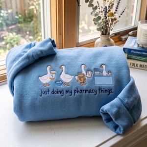 Könnte beinhalten: Ein hellblaues Sweatshirt mit einer Stickerei von vier weißen Gänsen, die pharmazeutische Aufgaben erledigen, darunter das Halten einer Arzneiflasche, einer Papiertüte und eines Klemmbretts, mit dem Text "just doing my pharmacy things."
