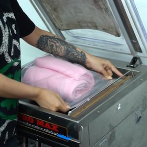 May include: A stainless steel vacuum sealer machine with a clear hinged lid, featuring a control panel with a red emergency stop button and a rotary dial. Inside the chamber, a folded pink textile item is placed within a clear plastic bag.