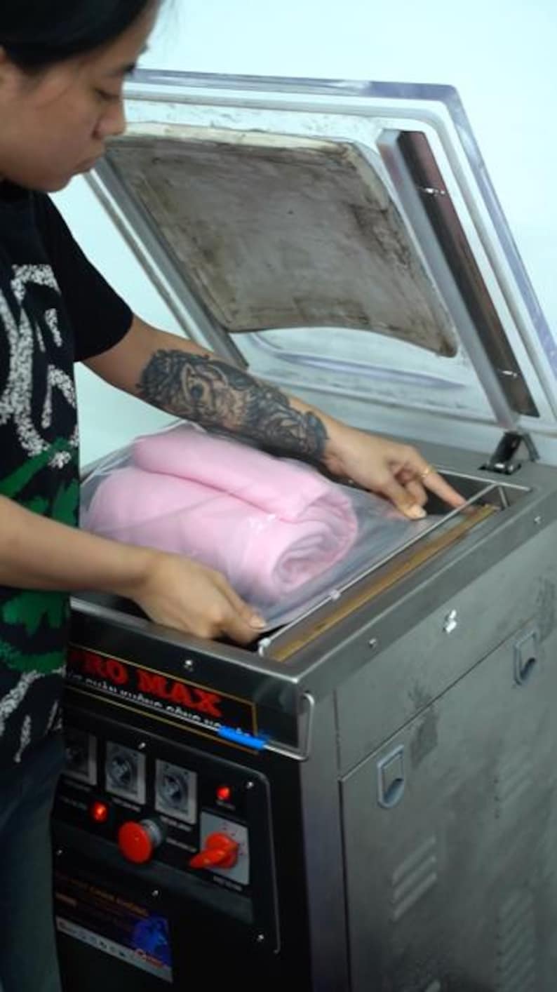 May include: A stainless steel vacuum sealer machine with a clear hinged lid, featuring a control panel with a red emergency stop button and a rotary dial. Inside the chamber, a folded pink textile item is placed within a clear plastic bag.
