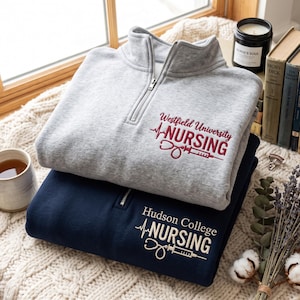 May include: A light gray quarter-zip sweatshirt with maroon embroidery reading Westfield University NURSING and a stethoscope and syringe icon, stacked on a navy blue quarter-zip sweatshirt with white embroidery reading Hudson College NURSING.