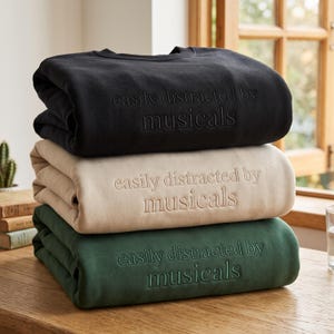 Könnte beinhalten: Ein Stapel aus drei Rundhals-Sweatshirts in Schwarz, Beige und Dunkelgrün. Jedes Sweatshirt ist auf der Brust mit dem Text "easily distracted by musicals" in passender Garnfarbe bestickt, was einen dezenten Ton-in-Ton-Effekt erzeugt.