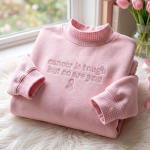 Op de afbeelding: Een lichtroze sweatshirt met een opstaande kraag en mouwboorden met een wit en roze gingham-patroon. De voorzijde is geborduurd met de tekst "cancer is tough but so are you" boven een klein roze symbool van een bewustwordingslint.