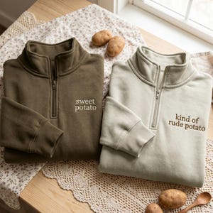 May include: Two quarter-zip sweatshirts with ribbed cuffs and collars. The dark olive sweatshirt features the embroidered text "sweet potato" and the light beige sweatshirt features the embroidered text "kind of rude potato".